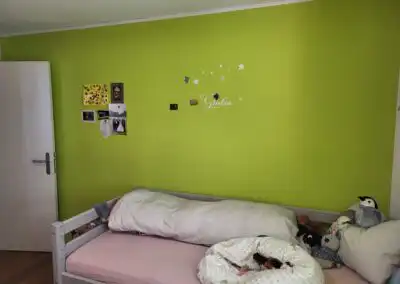 Kinderzimmer mit grüner Wand, weißen Sternen, Bett mit Kuscheltieren und persönlichen Erinnerungsfotos dekoriert.