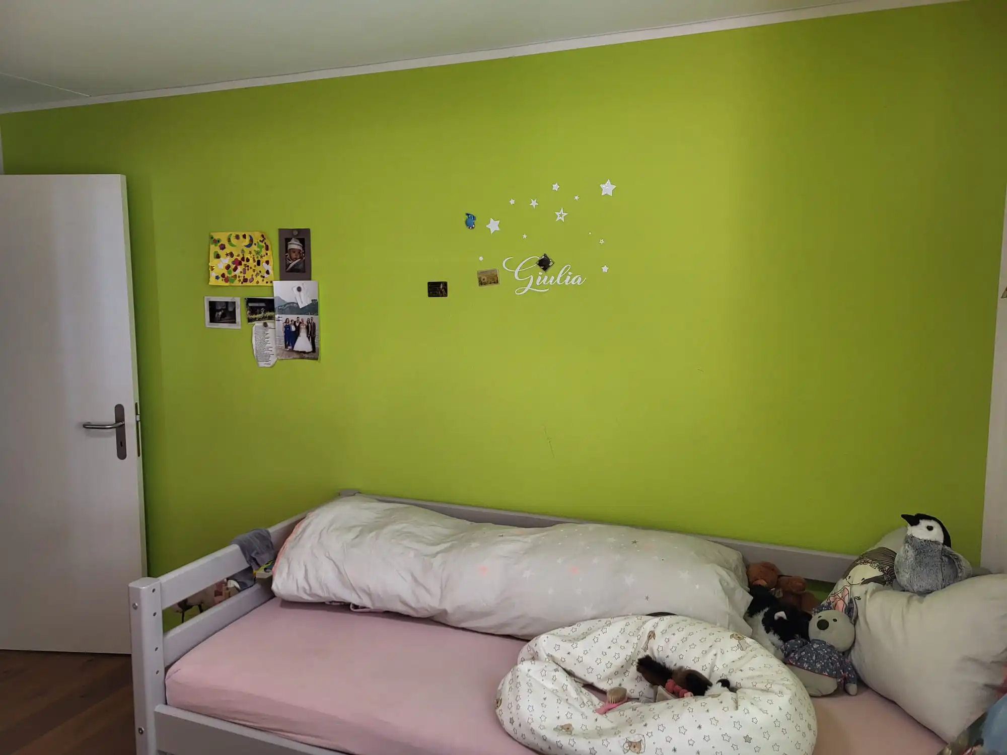 Kinderzimmer mit grüner Wand, weißen Sternen, Bett mit Kuscheltieren und persönlichen Erinnerungsfotos dekoriert.