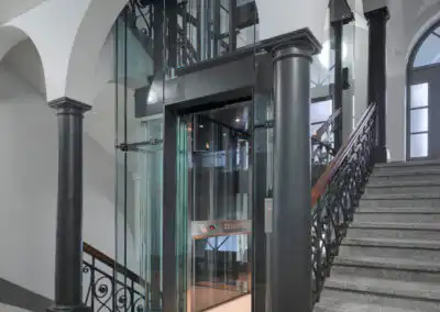 Moderner Glasaufzug in historischem Treppenhaus mit Bögen und schwarzem Geländer. Elegante Kombination aus Alt und Neu.