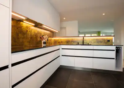 Moderne weiße Küche mit schwarzer Arbeitsplatte und goldener Rückwand, stilvolle Beleuchtung, minimalistisches Design.
