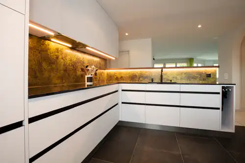 Moderne weiße Küche mit schwarzer Arbeitsplatte und goldener Rückwand, stilvolle Beleuchtung, minimalistisches Design.