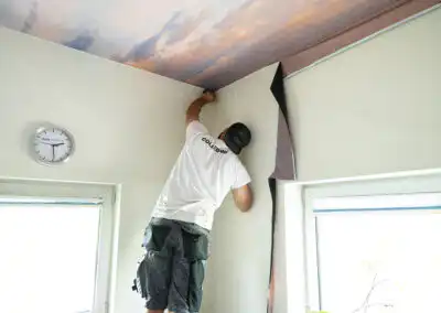 Handwerker montiert eine Fototapete an der Decke in einem Wohnraum, dabei steht eine Uhr an der Wand im Blick.