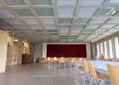 Leerer Konferenzsaal mit langer Tafel, Holzstühlen und rotem Vorhang, Fensterfront mit hellem Tageslicht.