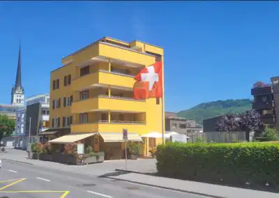 Gelbes Gebäude mit Schweizer Flagge, im Hintergrund eine Kirche, an einem sonnigen Tag in einem Stadtgebiet.