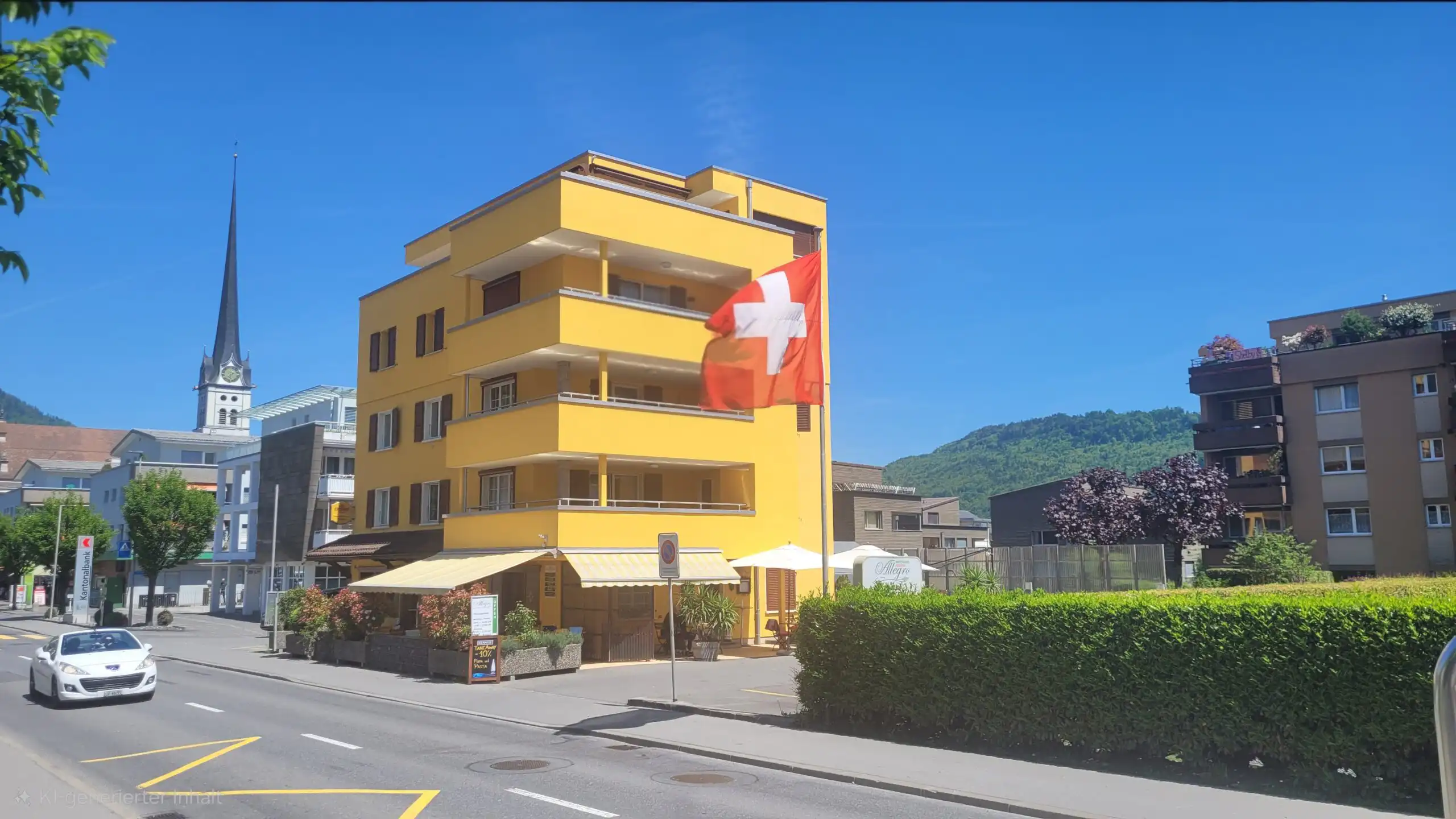 Gelbes Gebäude mit Schweizer Flagge, im Hintergrund eine Kirche, an einem sonnigen Tag in einem Stadtgebiet.