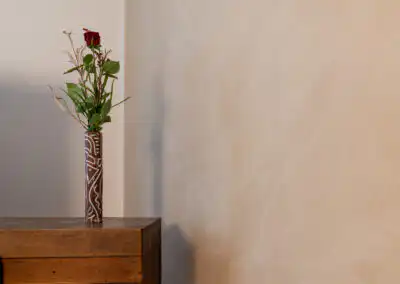 Rote Rose in verziertem Holzvase auf Holztisch vor beiger Wand, minimalistisch dekoriert.