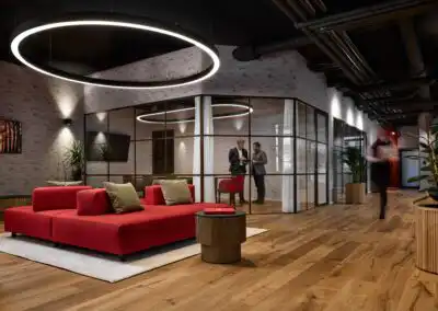Moderne Büro-Lounge mit rotem Sofa, Holzfußboden, stilvoller Beleuchtung und zwei Personen im Gespräch.