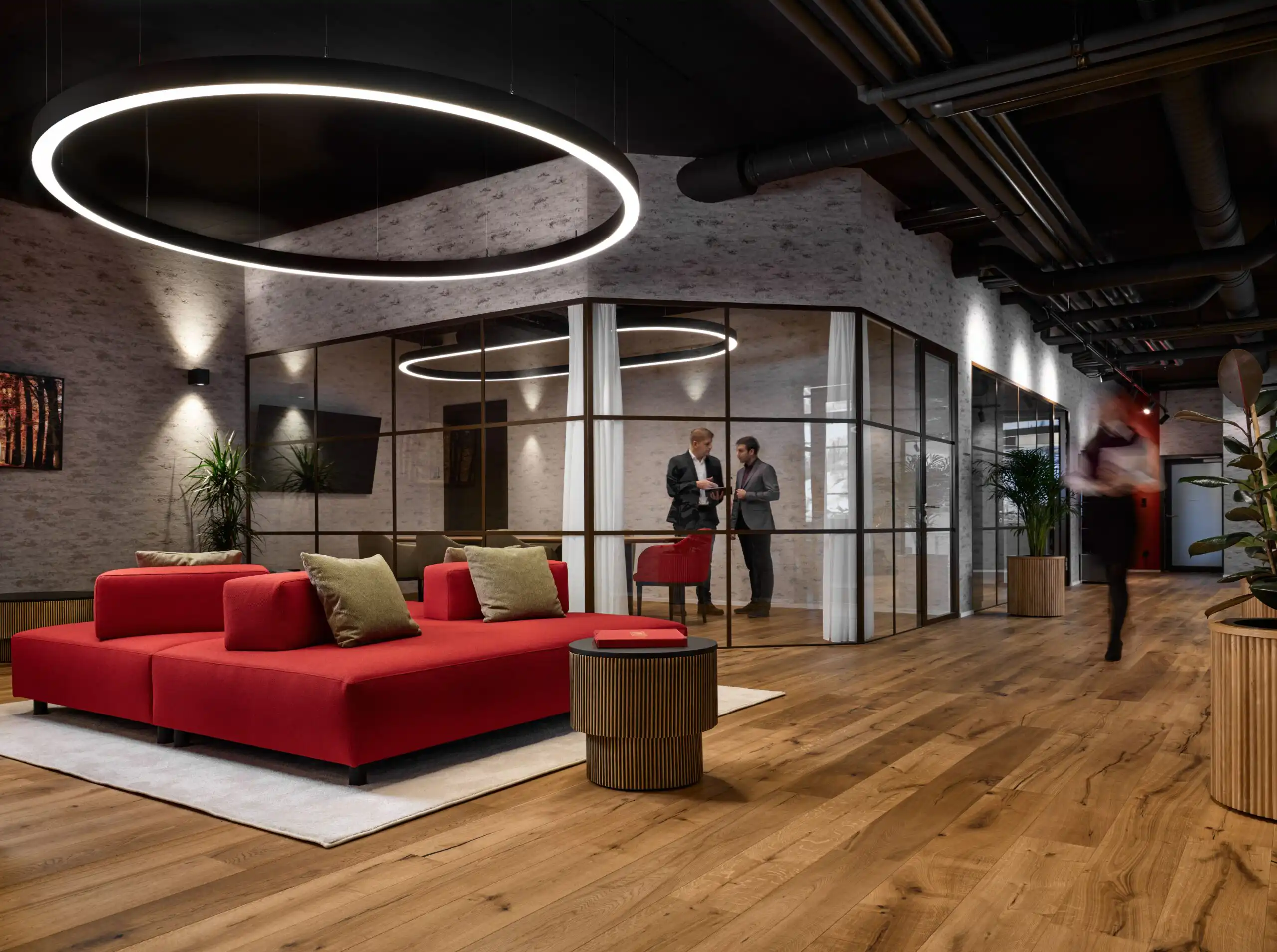 Moderne Büro-Lounge mit rotem Sofa, Holzfußboden, stilvoller Beleuchtung und zwei Personen im Gespräch.