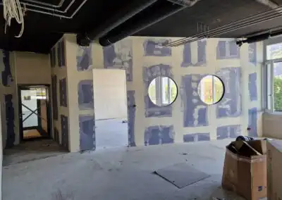Renovierter Raum mit runden Fenstern, freiliegendem Rohrsystem und Baustellenmaterialien auf dem Boden.