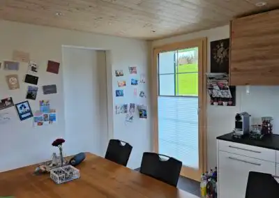 Moderner Essbereich mit Holztisch, Wanddekoration und Küchenzeile. Heller Raum mit Fensterblick ins Grüne.