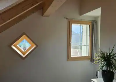 Dachgeschosszimmer mit Holzbalken, zwei Fenstern und Zimmerpflanze, Aussicht auf Berge.
