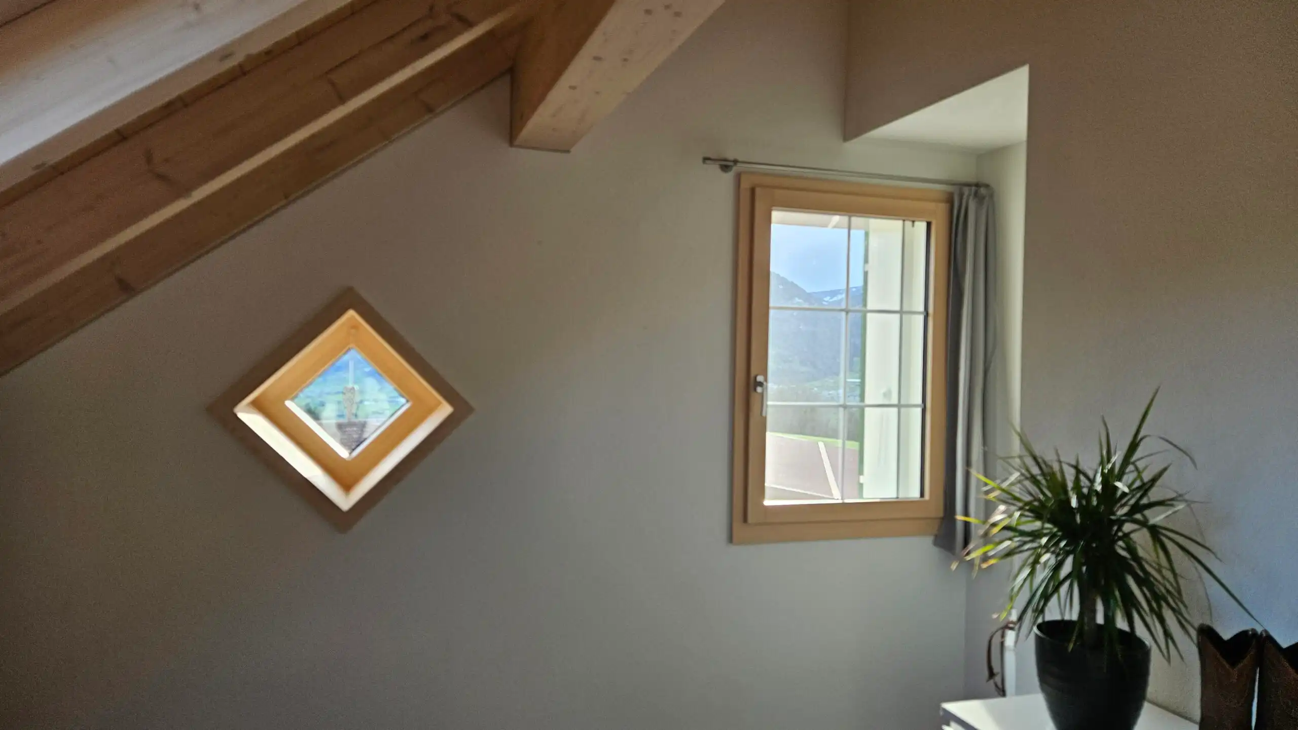 Dachgeschosszimmer mit Holzbalken, zwei Fenstern und Zimmerpflanze, Aussicht auf Berge.