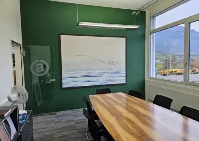 Moderner Besprechungsraum mit großem Holztisch, Kunstwerk an der grünen Wand und Blick auf Landschaft durch Fenster.