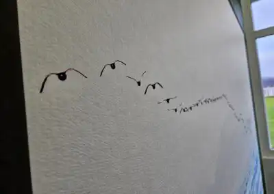 Minimalistisches Kunstwerk: Vogelschwarm auf Leinwand fliegt in Richtung Himmel, mit Fensteraussicht in der Ecke.