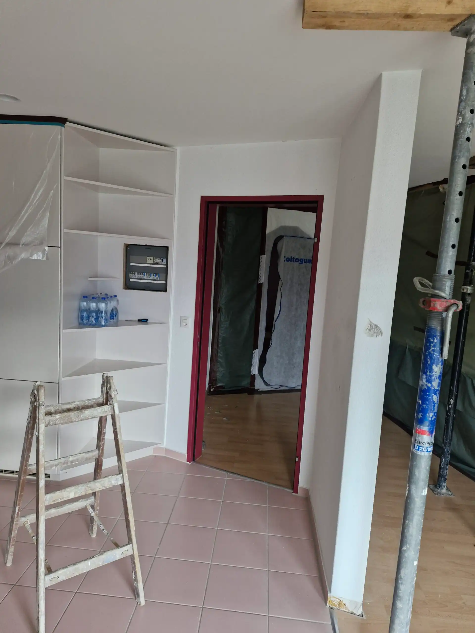 Renovierung in einem Wohnraum mit Leiter, Fliesenboden und Türrahmen in Rot. Werkzeuge für Innenausbau sichtbar.