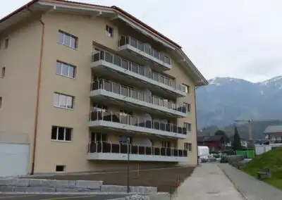 Mehrfamilienhaus mit Balkonen vor Berglandschaft, wolkiger Himmel im Hintergrund.