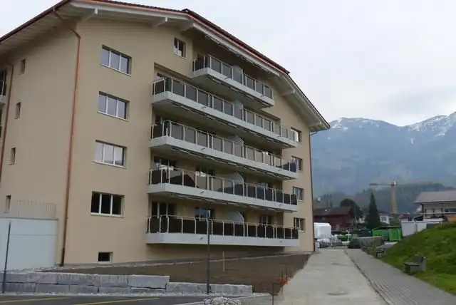 Mehrfamilienhaus mit Balkonen vor Berglandschaft, wolkiger Himmel im Hintergrund.