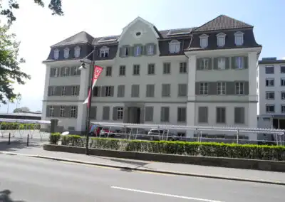 Historisches Gebäude mit Satteldach und Fensterläden, dekoriert mit Fahnen an einer ruhigen Straße bei Sonnenschein.