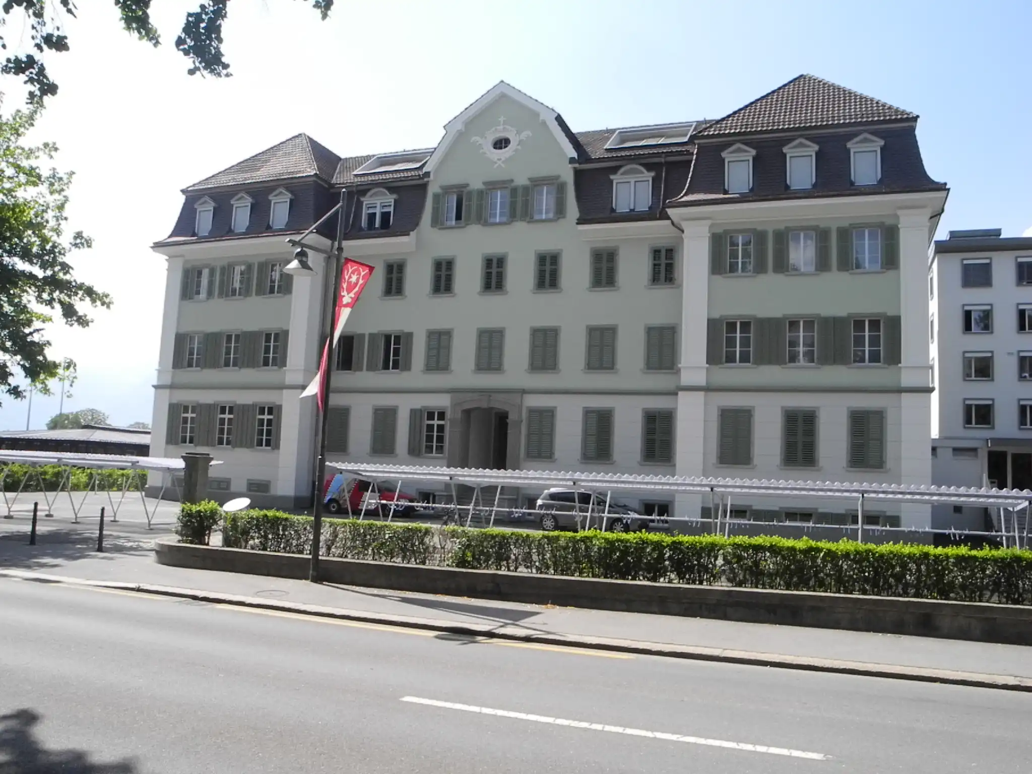 Historisches Gebäude mit Satteldach und Fensterläden, dekoriert mit Fahnen an einer ruhigen Straße bei Sonnenschein.