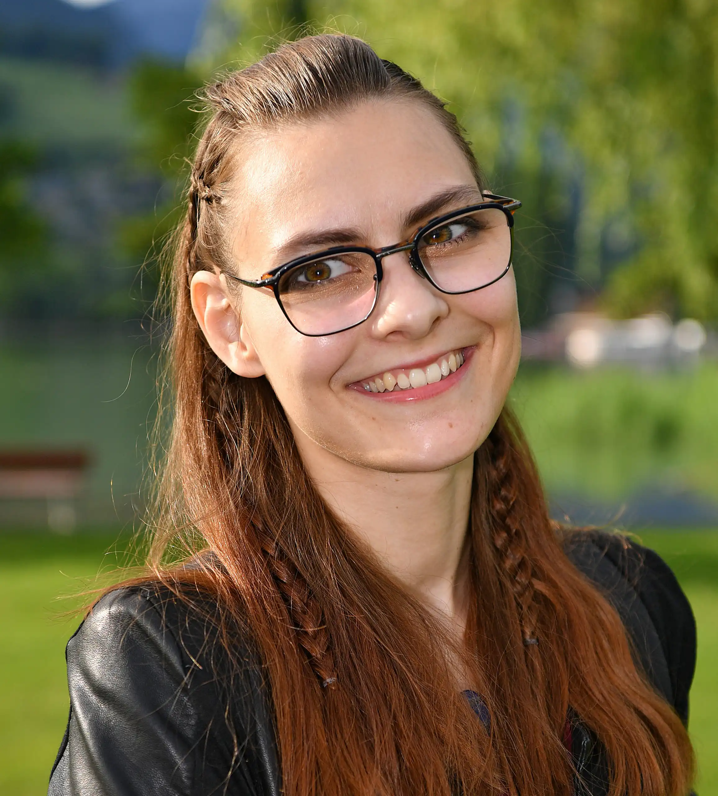 Frau mit Brille und geflochtenem Haar lächelt im Freien, grüne Landschaft im Hintergrund.