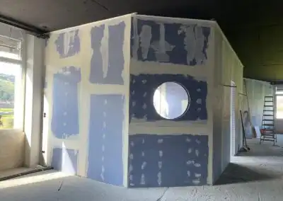 Unfertiger Innenausbau mit Trockenbauwand und rundem Fenster, Leiter im Hintergrund, helles Tageslicht.