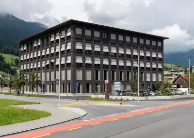 Modernes Bürogebäude vor Alpen, umgeben von Grünflächen und Straßen, mit bewölktem Himmel im Hintergrund.