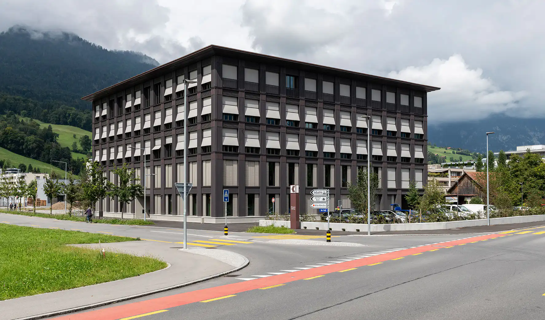 Modernes Bürogebäude vor Alpen, umgeben von Grünflächen und Straßen, mit bewölktem Himmel im Hintergrund.